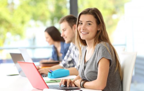 Happy student posing in a classroom looking at you with an unfocused window in the background